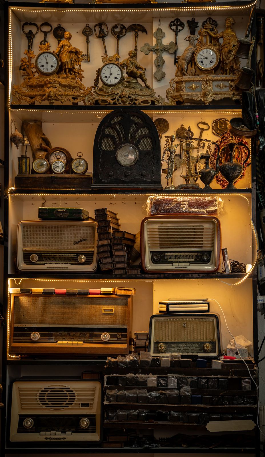 Warmly lit shelving of antique tube radios, gilded mantel clocks with figures, ornate keys and small brass ornaments