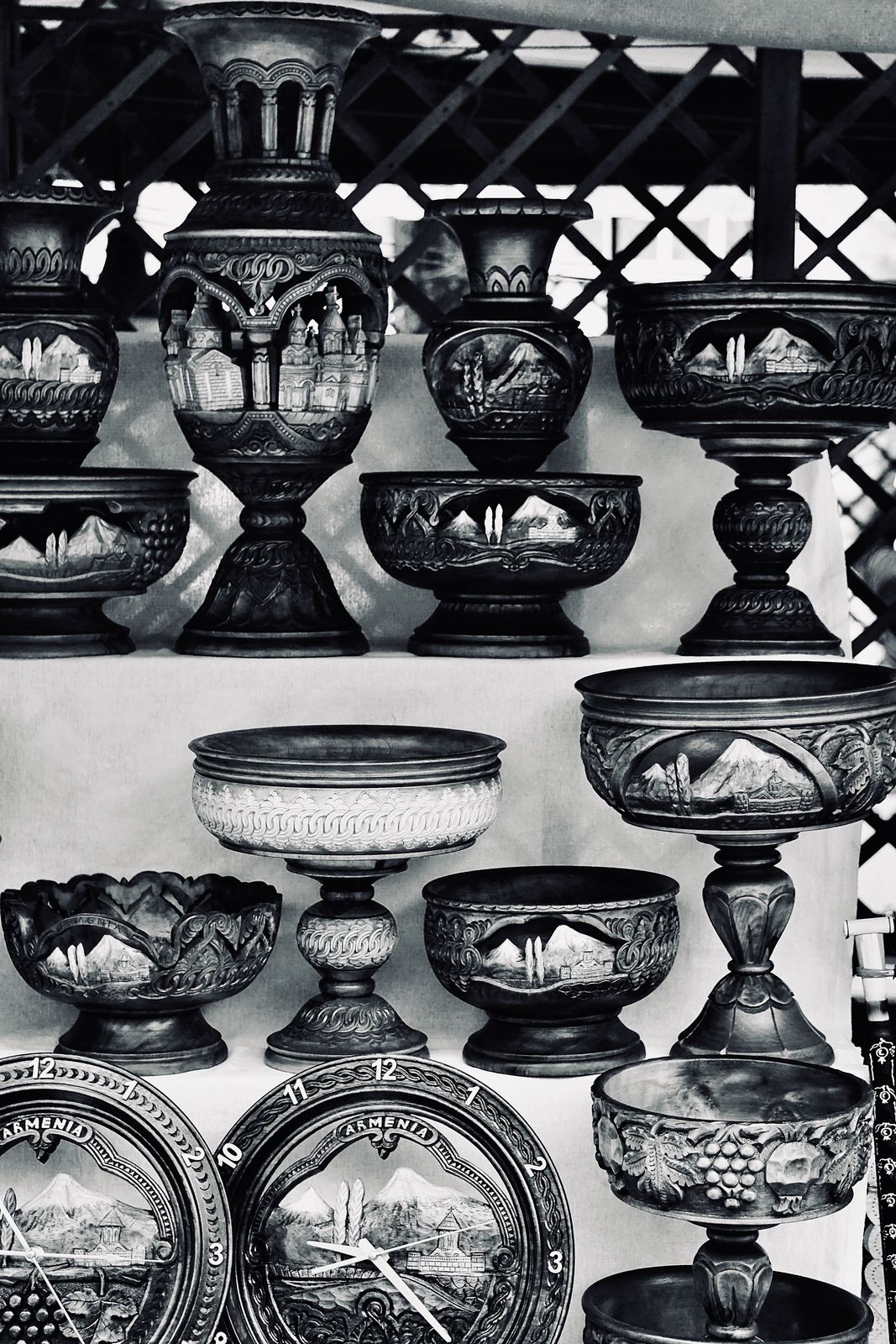 Black-and-white photo of hand-carved wooden vases and footed bowls with mountain and church scenes, arranged on tiered shelves