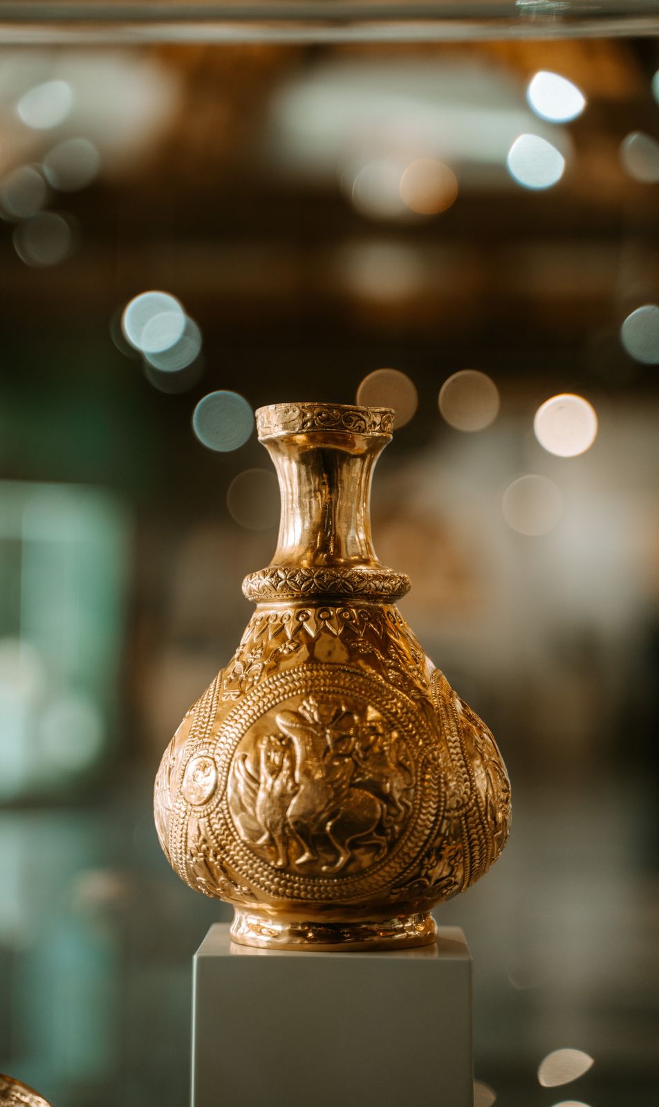 Gilded vase embossed with a rider medallion and beaded borders, displayed on a white plinth with soft background lights