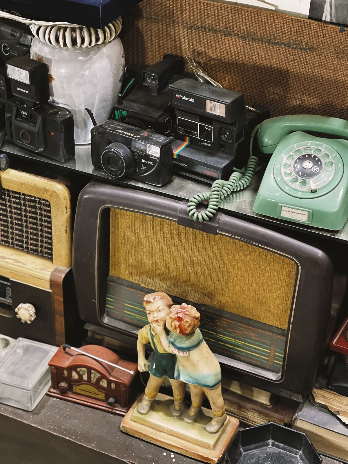Vintage instant and compact film cameras on a shelf above a mid-century radio, green rotary telephone and painted figurine of two children