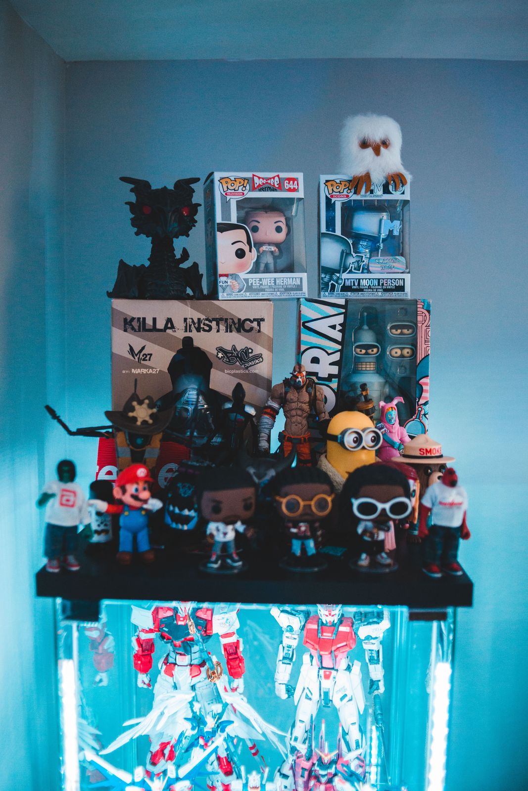 Stacked display of boxed vinyl figures, plush owl and assorted small figures above a lit cabinet of white and red mecha model kits